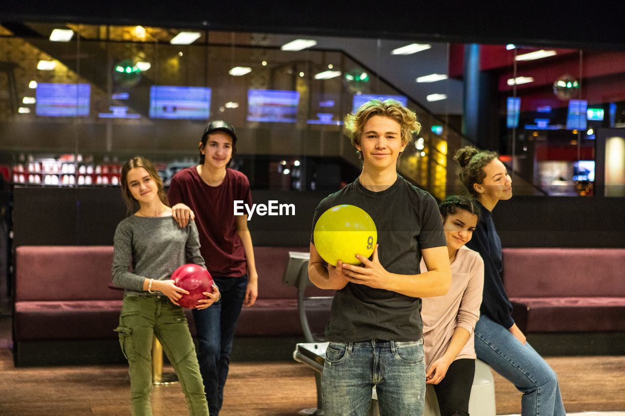 Portrait of smiling teenage boy holding ball while standing against friends at bowling alley