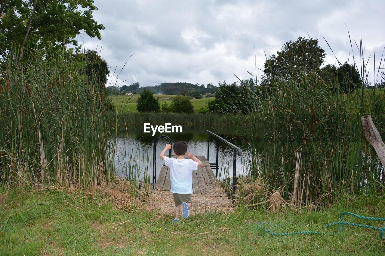 Rear view of boy walking on grassy field leading towards pond