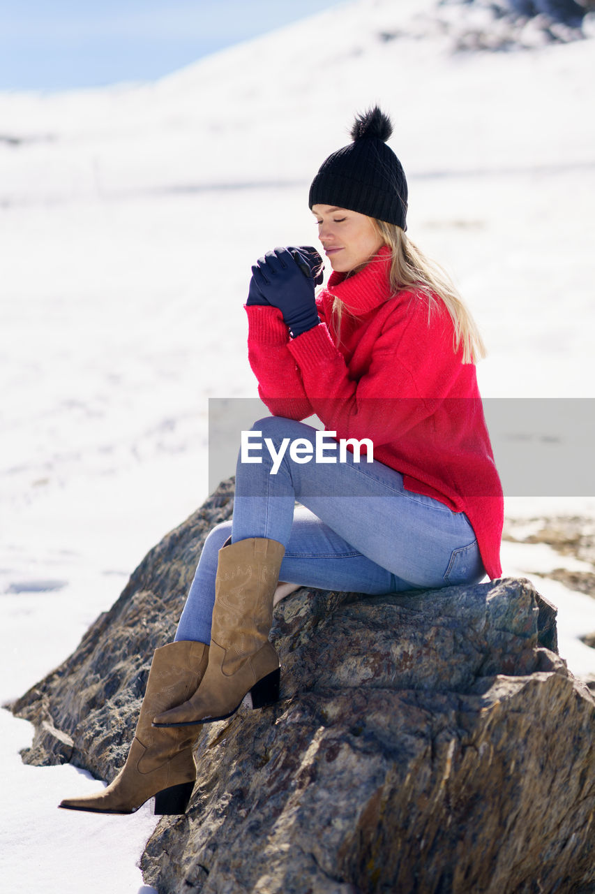 Full length of young woman sitting on rock on snow covered land