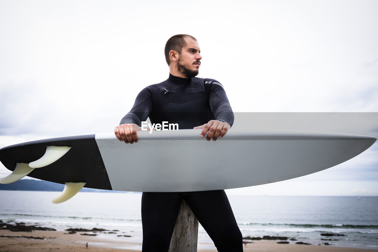 Man holding surfboard standing at beach against sky