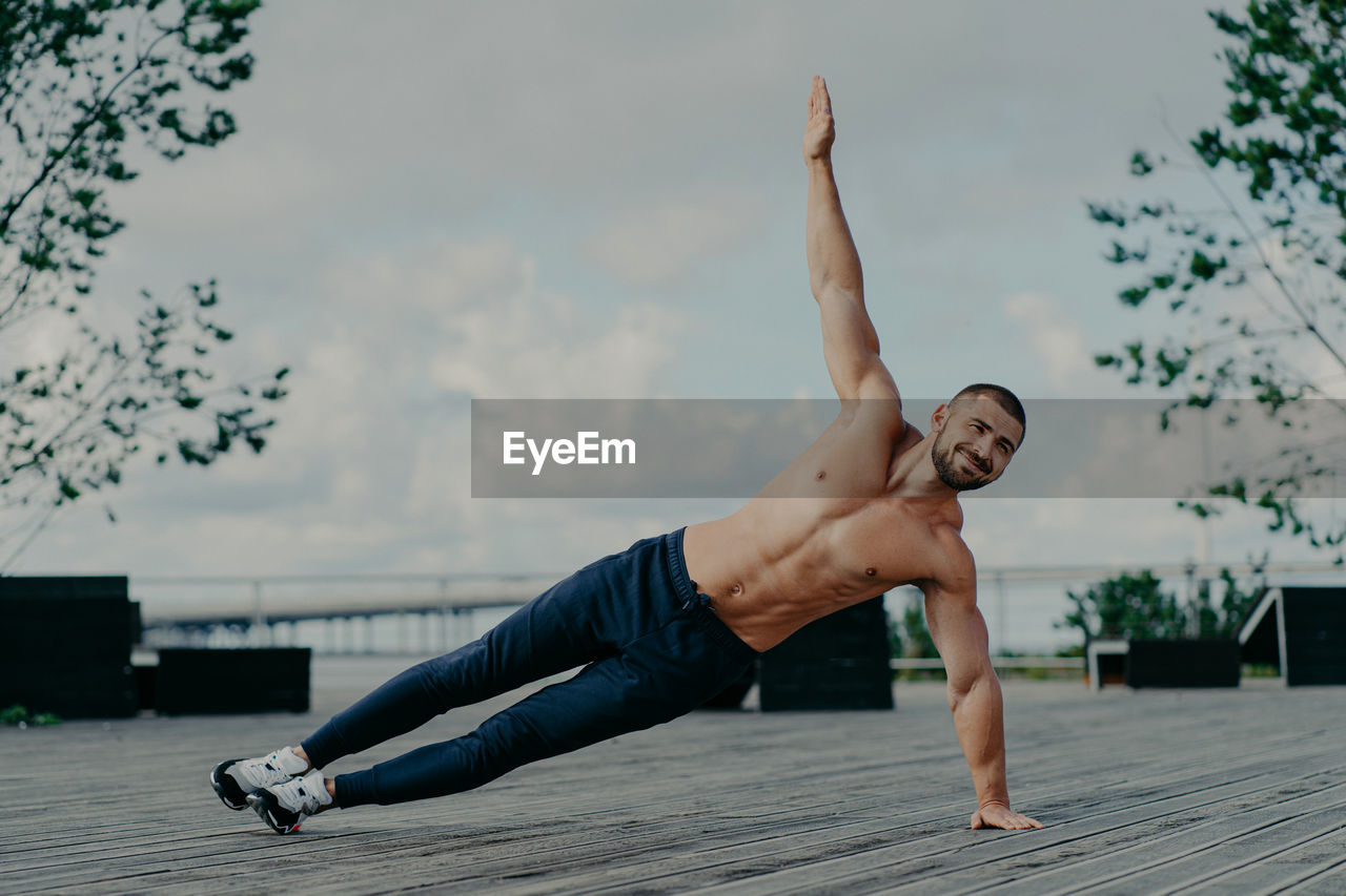 Full length of shirtless man with arms raised against sky