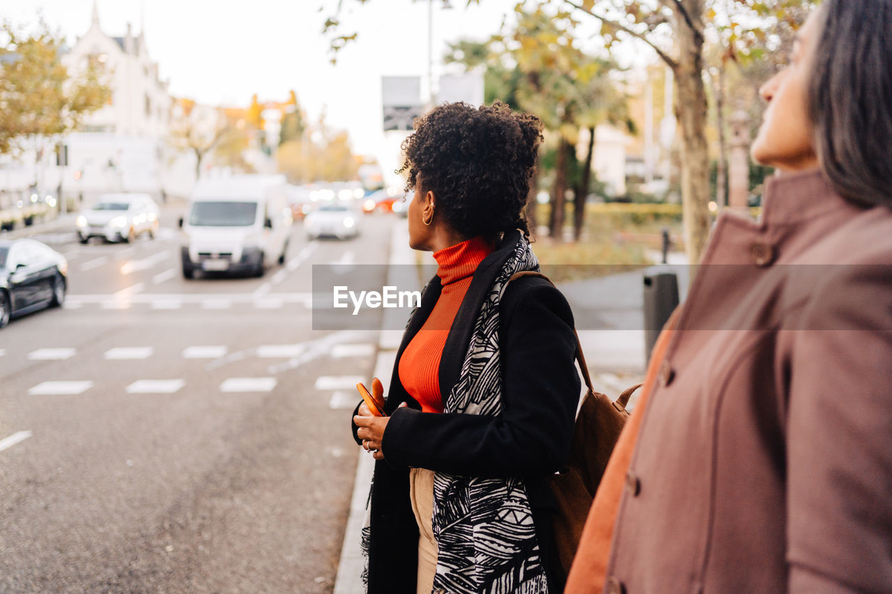 Side view of selective focus of ethnic female and hispanic lady in outerwear looking away while standing near asphalt road in city