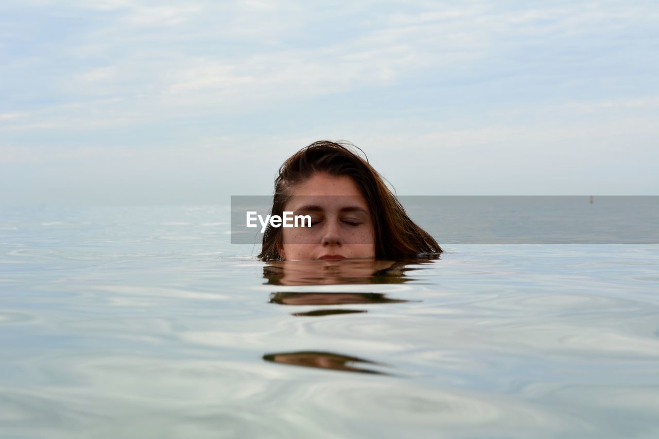 Close-up of young woman with eyes closed swimming in sea against cloudy sky