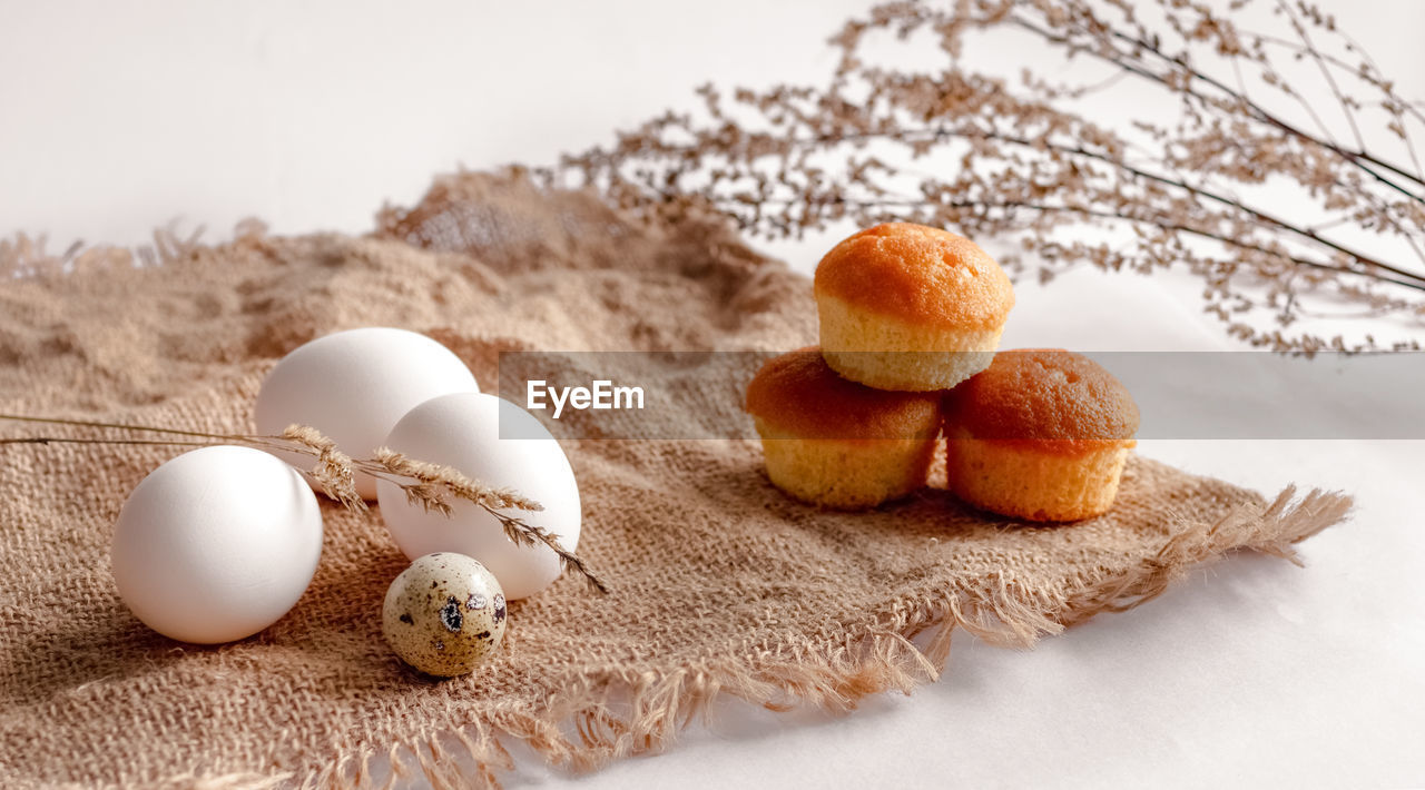 White chicken eggs, a quail egg, stems of dried grass and three cupcakes on a burlap bag