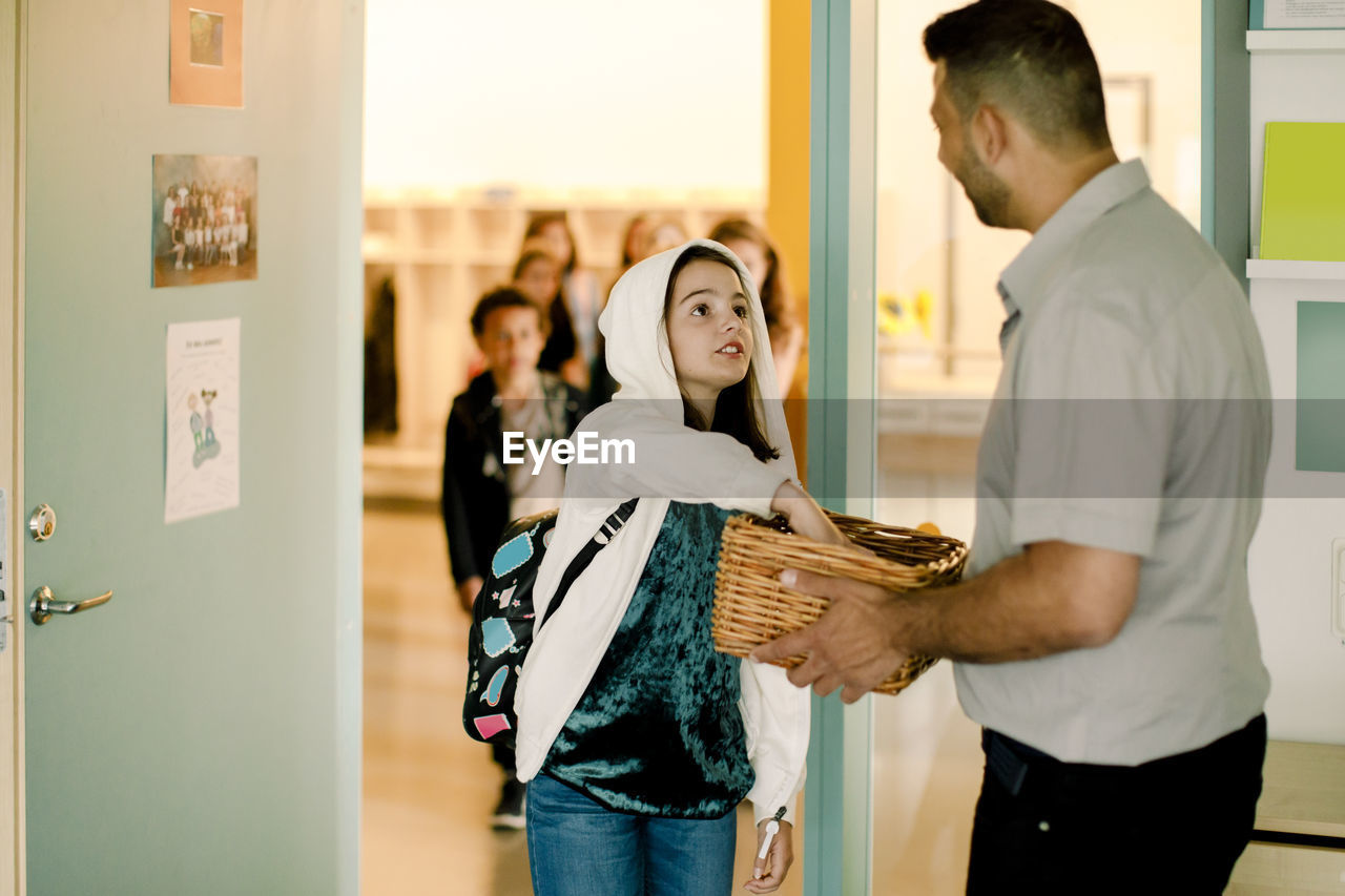 Girl in hooded shirt keeping phone while male teacher holding basket in classroom