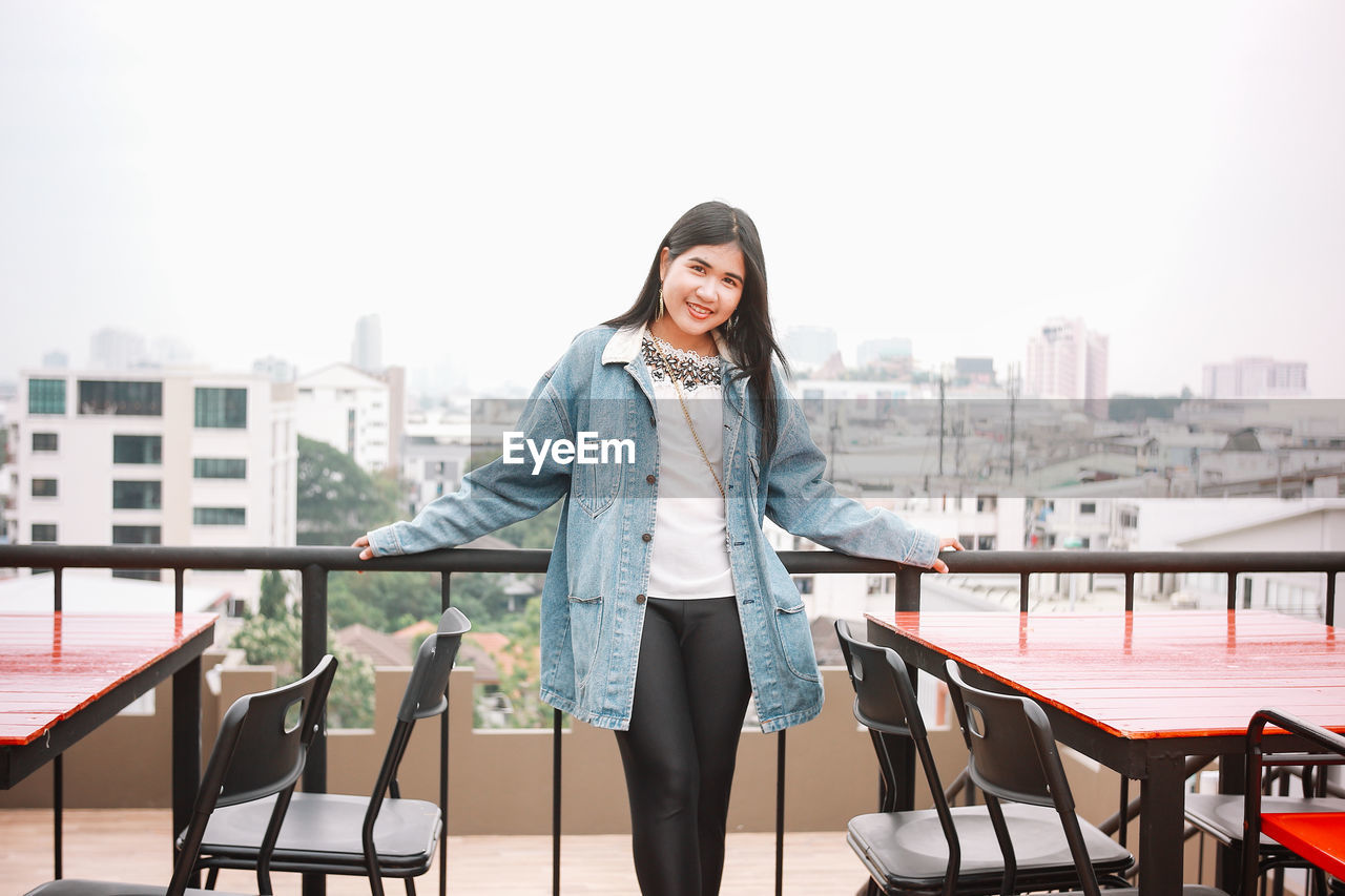 Portrait of young woman standing by railing against clear sky in city