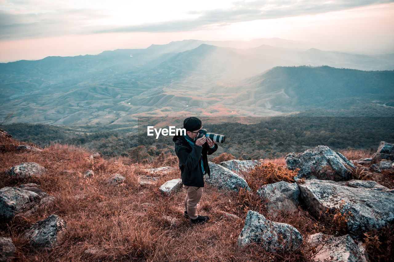 Man photographing through camera while standing on mountain against sky during sunset