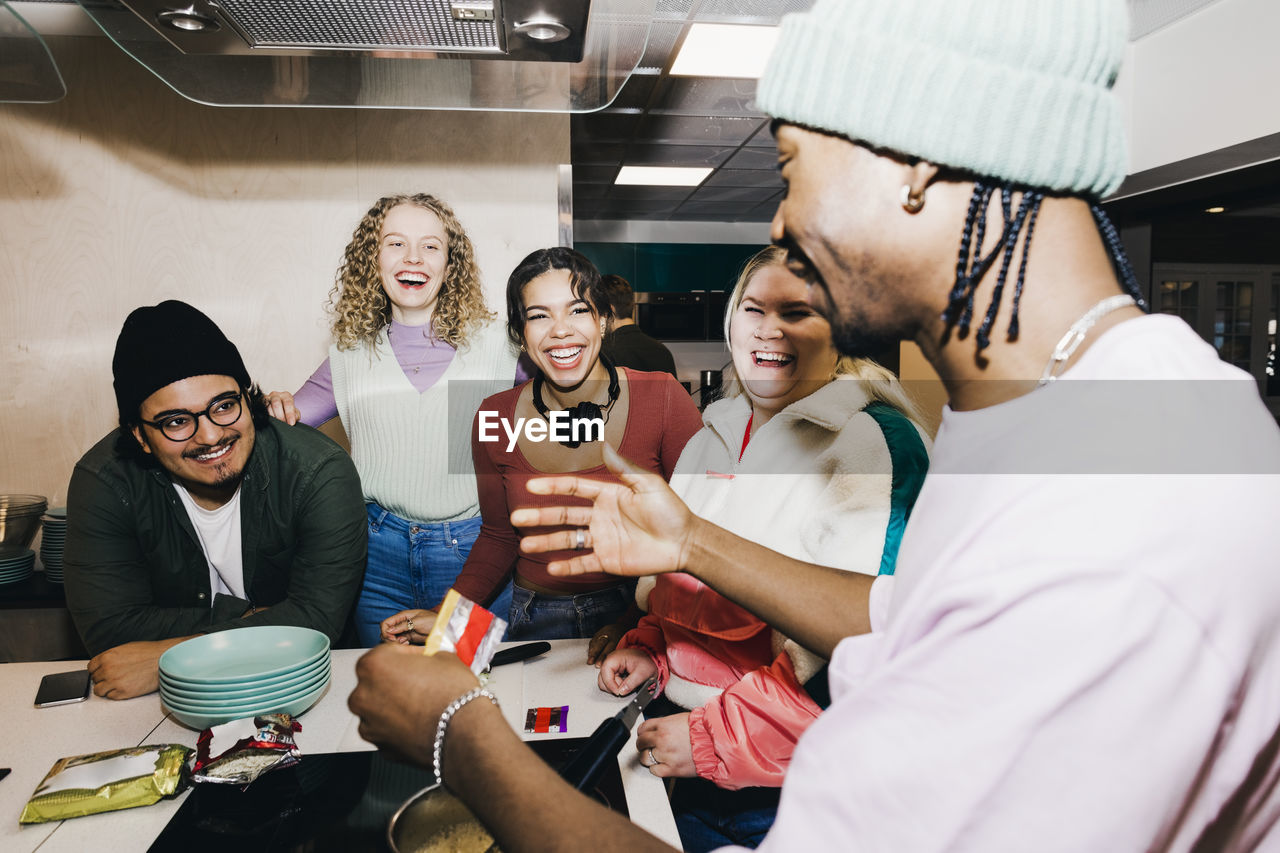 Happy multiracial young man and women looking at male friend preparing food in college dorm