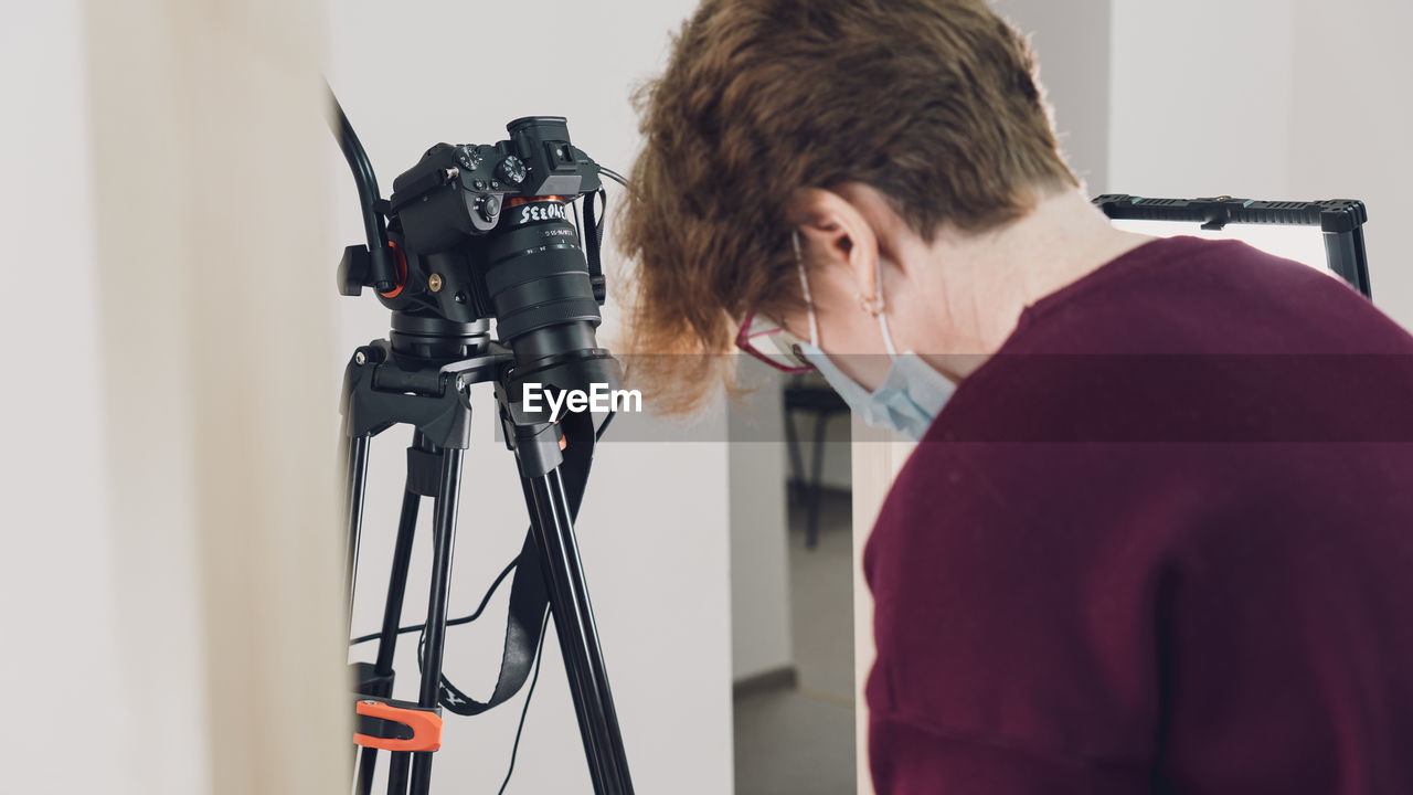 tripod, adult, indoors, one person, technology, occupation, studio, camera, arts culture and entertainment, filming, clothing, working, young adult, filmmaking, professional occupation, creativity, person, communication, waist up