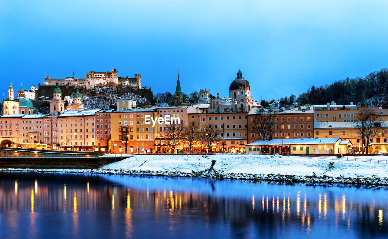 BUILDINGS BY RIVER AGAINST BLUE SKY IN WINTER
