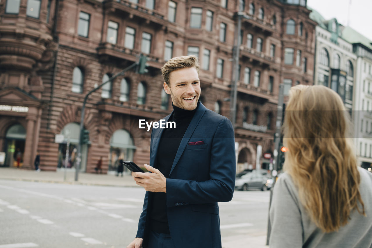 Smiling businessman with phone looking at female coworker while standing in city