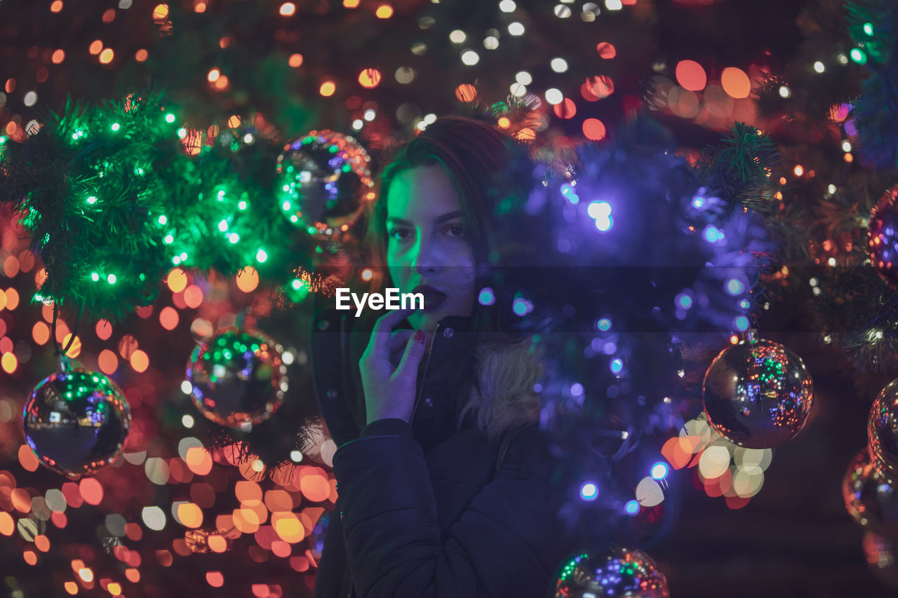 Thoughtful young woman standing amidst illuminated christmas tree at night