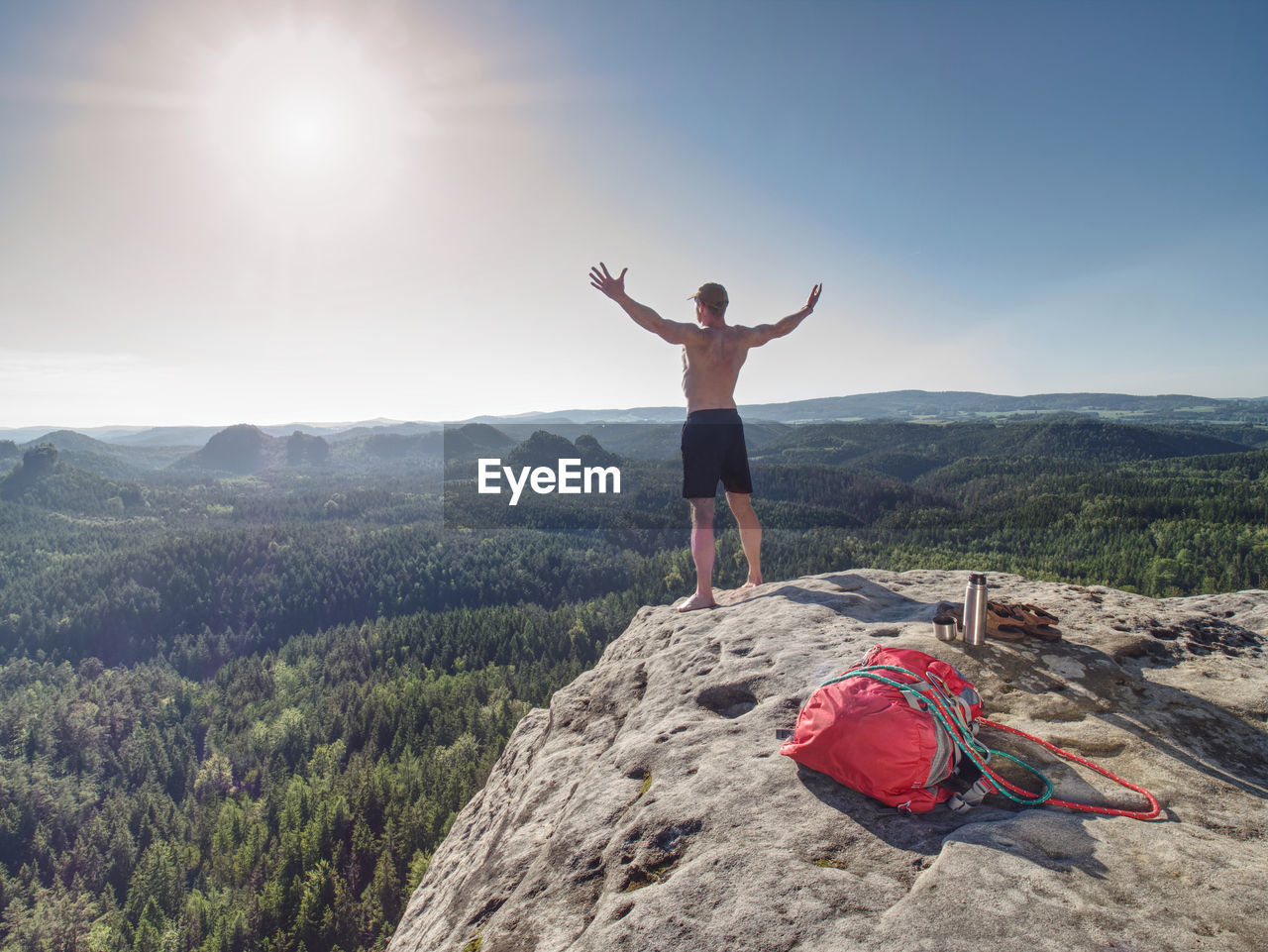 Shirtless sportsman take off backpack and rope on top of mountain and raised arms to greeting nature