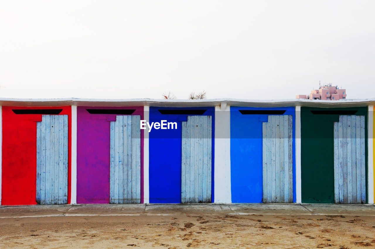 beach, architecture, land, blue, no people, sky, sand, beach hut, built structure, nature, day, wall, hut, building exterior, in a row, multi colored, outdoors, clear sky, side by side, building, copy space, wood, water