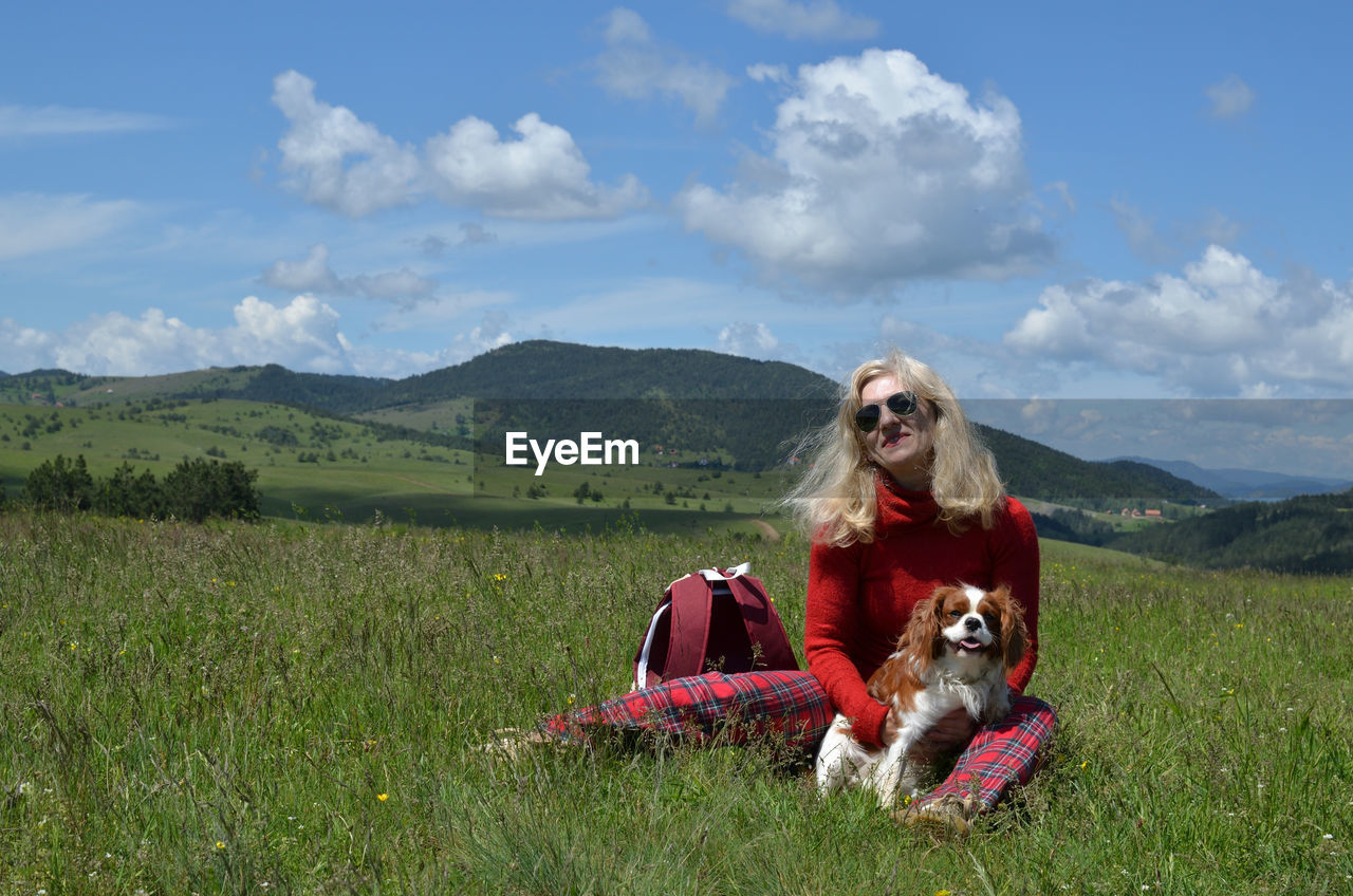 Happy blonde woman in red with her dog cavalier king charles spaniel on a meadow