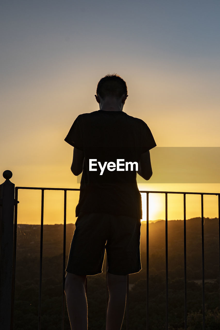 REAR VIEW OF SILHOUETTE BOY STANDING AGAINST SKY DURING SUNSET
