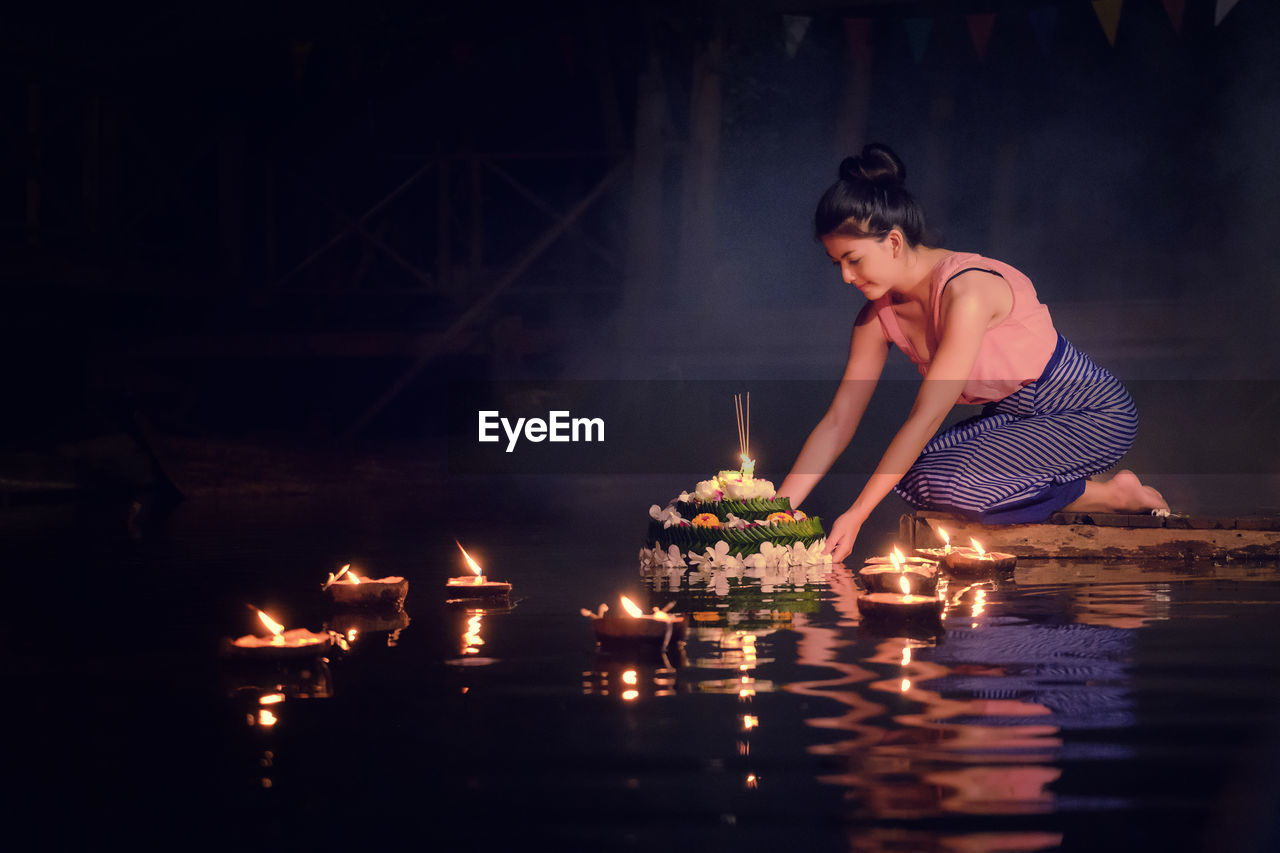 Young woman in traditional clothing kneeling by lake at night