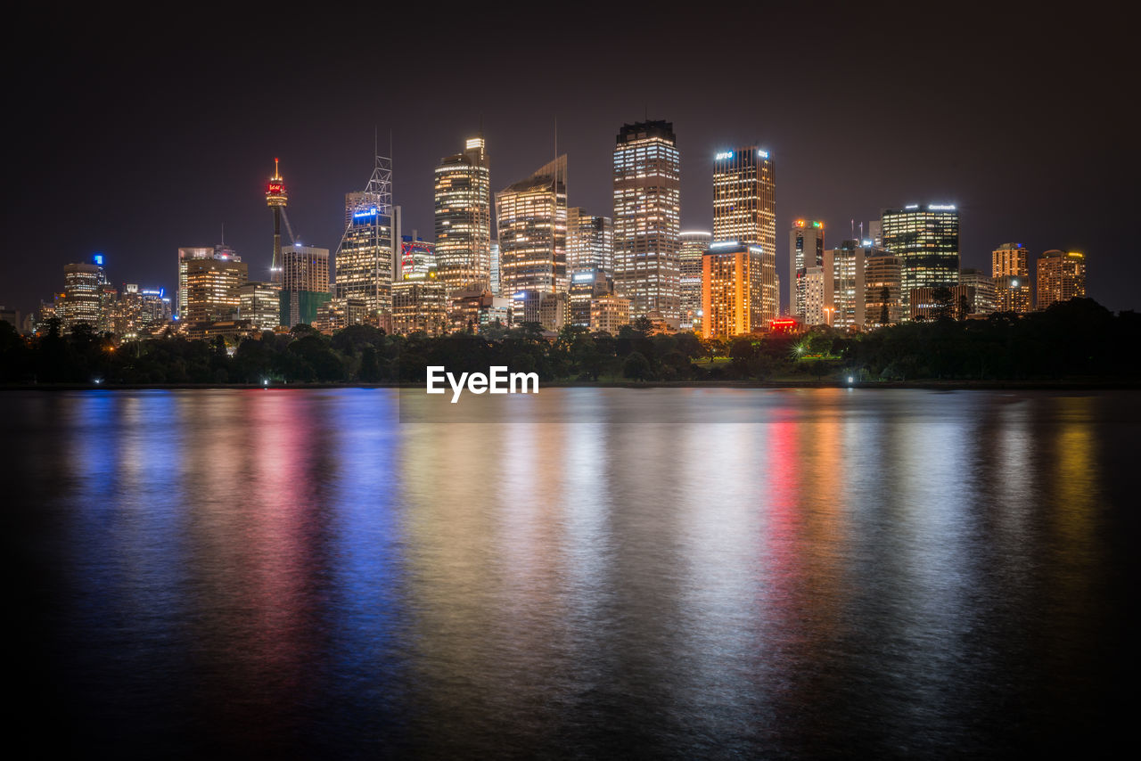 Illuminated buildings by river against sky at night