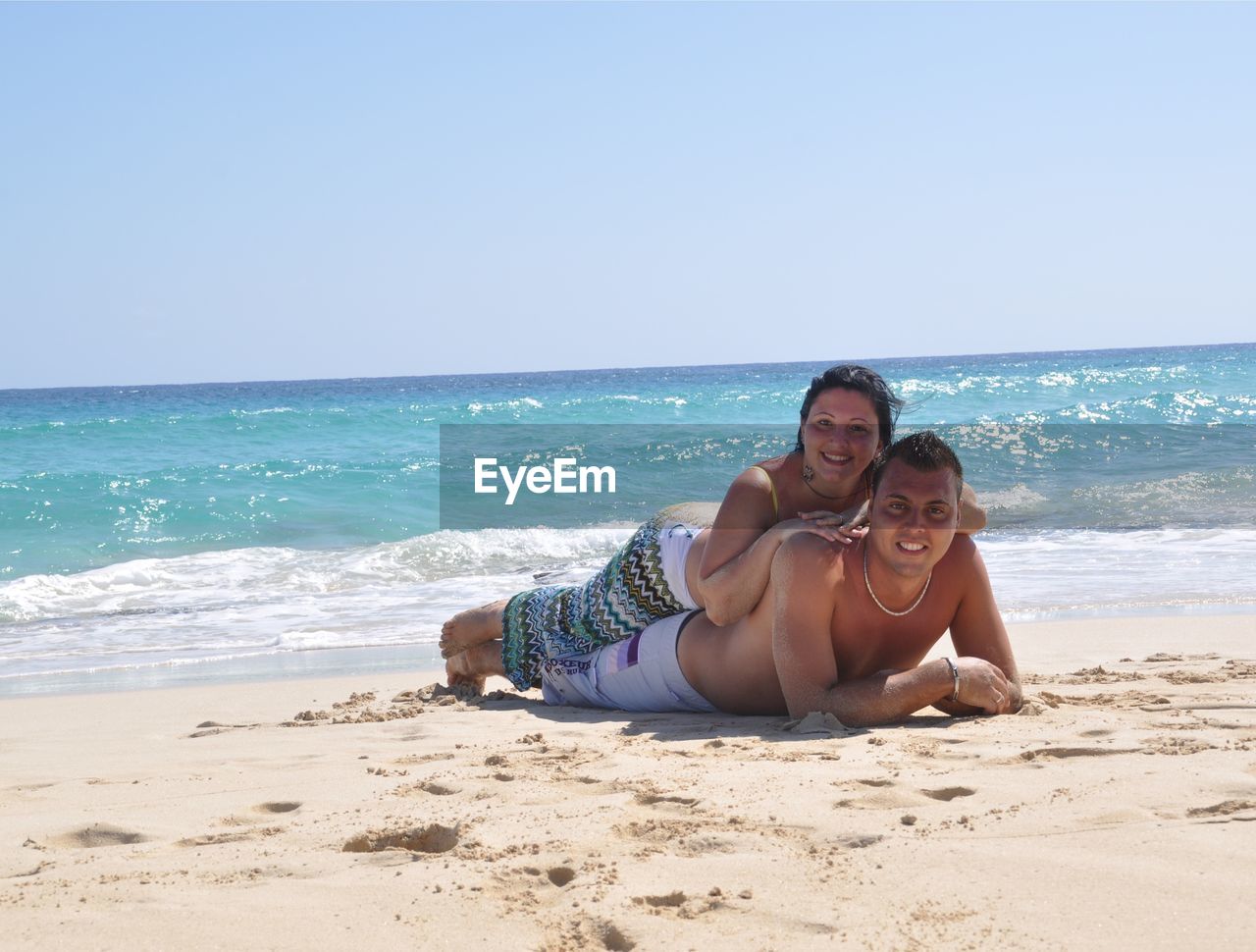 Portrait of smiling couple lying at beach against clear sky during sunny day