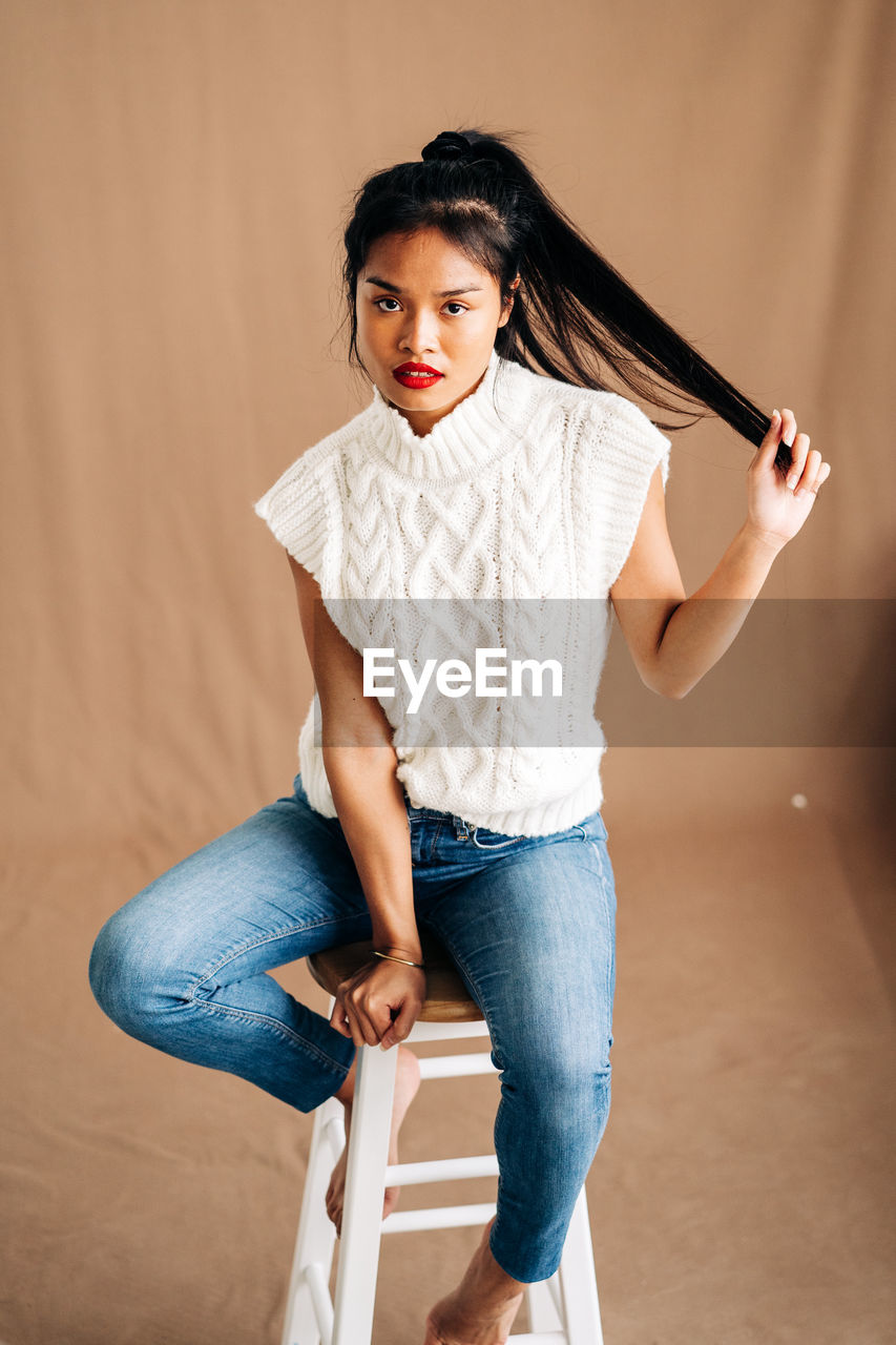 Confident young hispanic lady wearing casual clothes sitting on wooden stool in studio