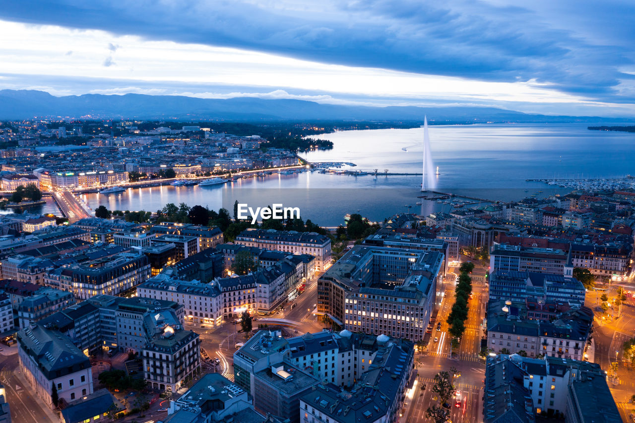 HIGH ANGLE VIEW OF ILLUMINATED CITY BY BUILDINGS AGAINST CLOUDY SKY