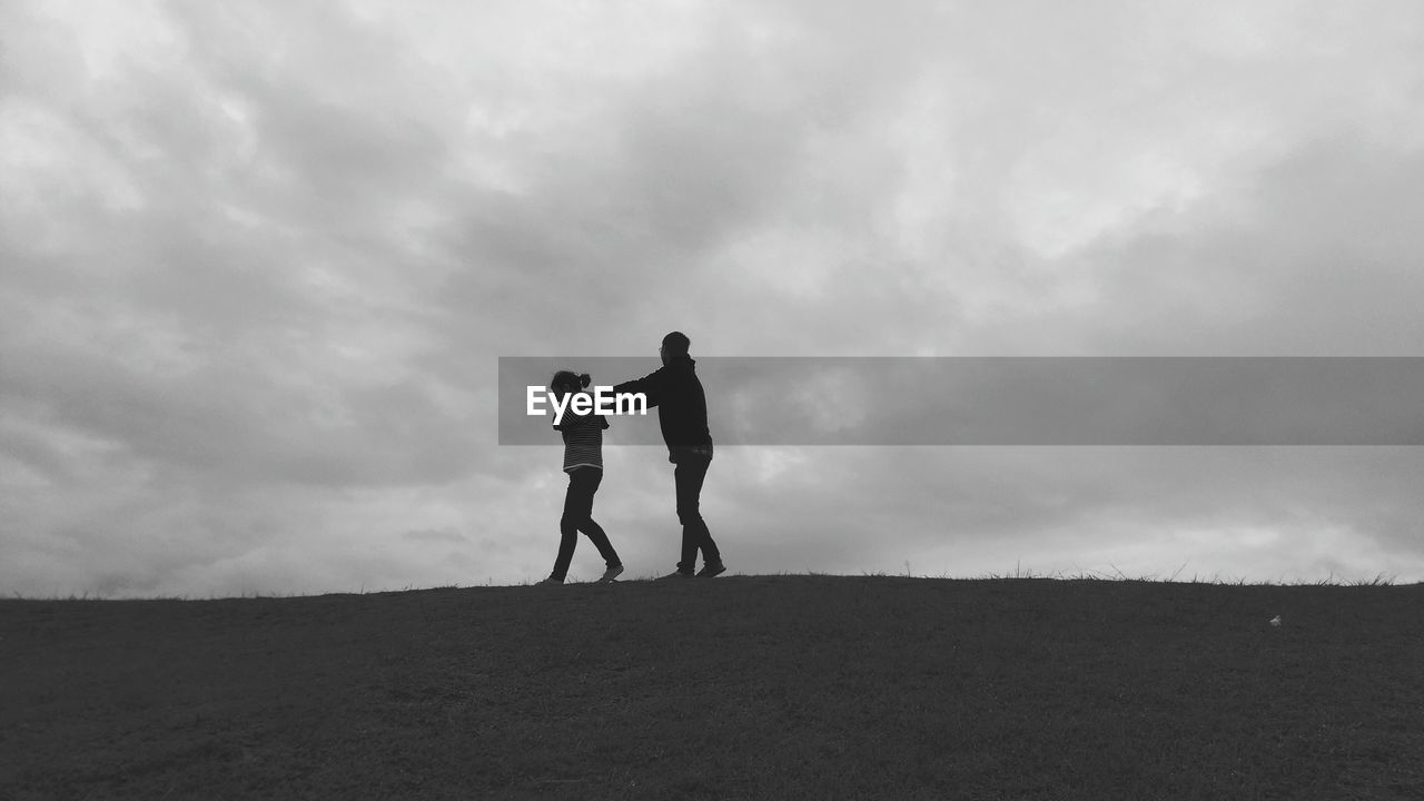 Low angle view of man and woman walking on hill against cloudy sky at dusk