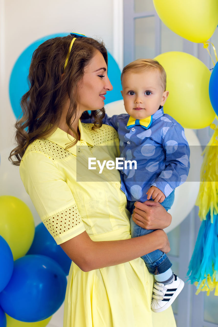 Happy mom in a yellow dress holds a young son in blue clothes , behind fly balls yellow, blue