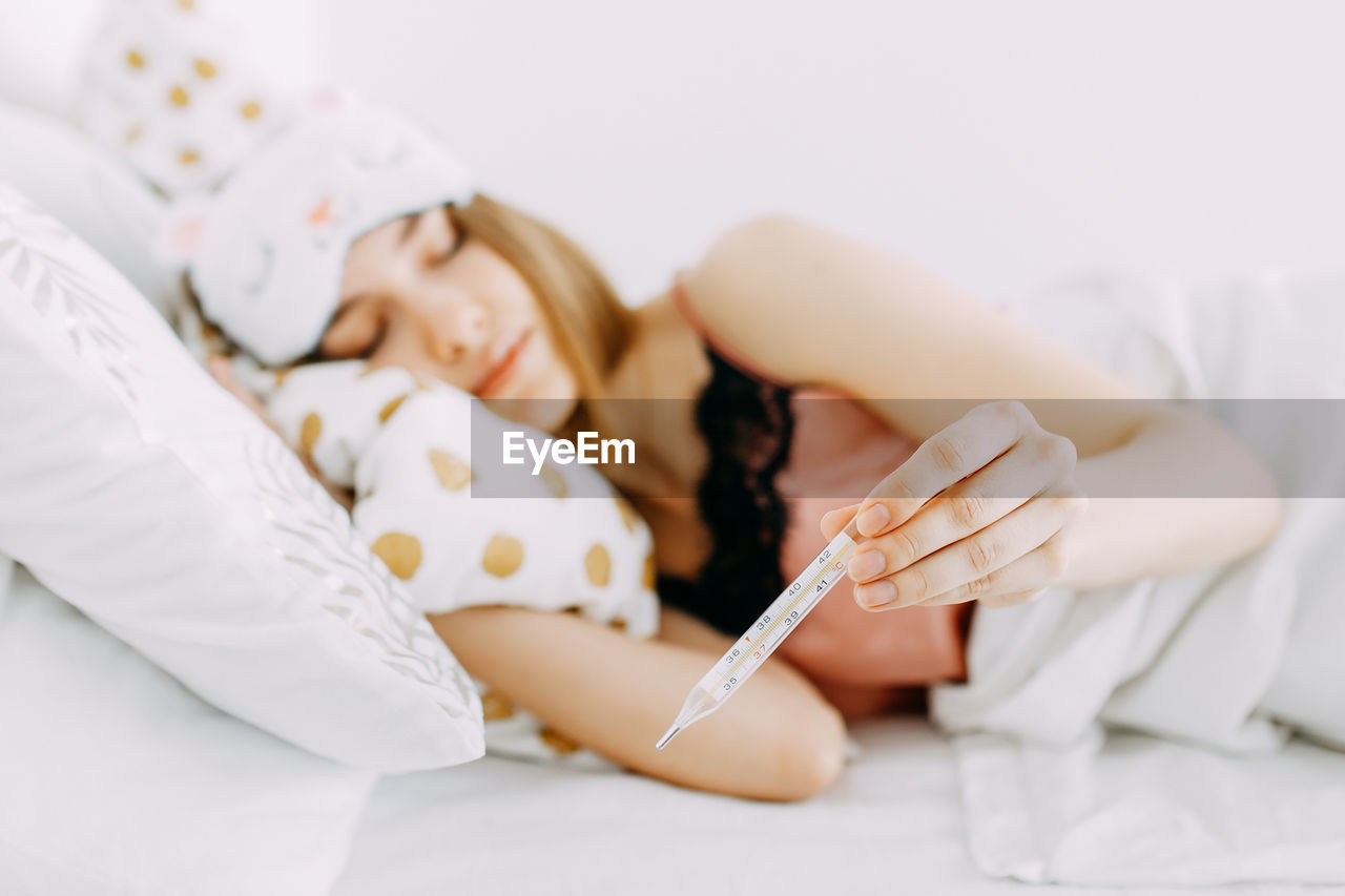 Young sick girl in pajamas lies with a temperature and a thermometer on the bed in the bedroom