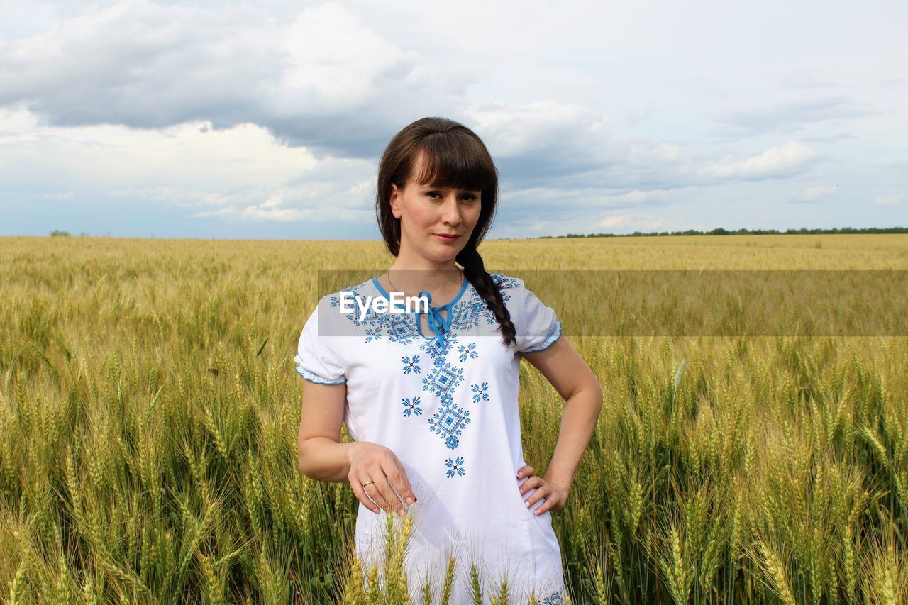 Portrait of young woman standing on field against cloudy sky