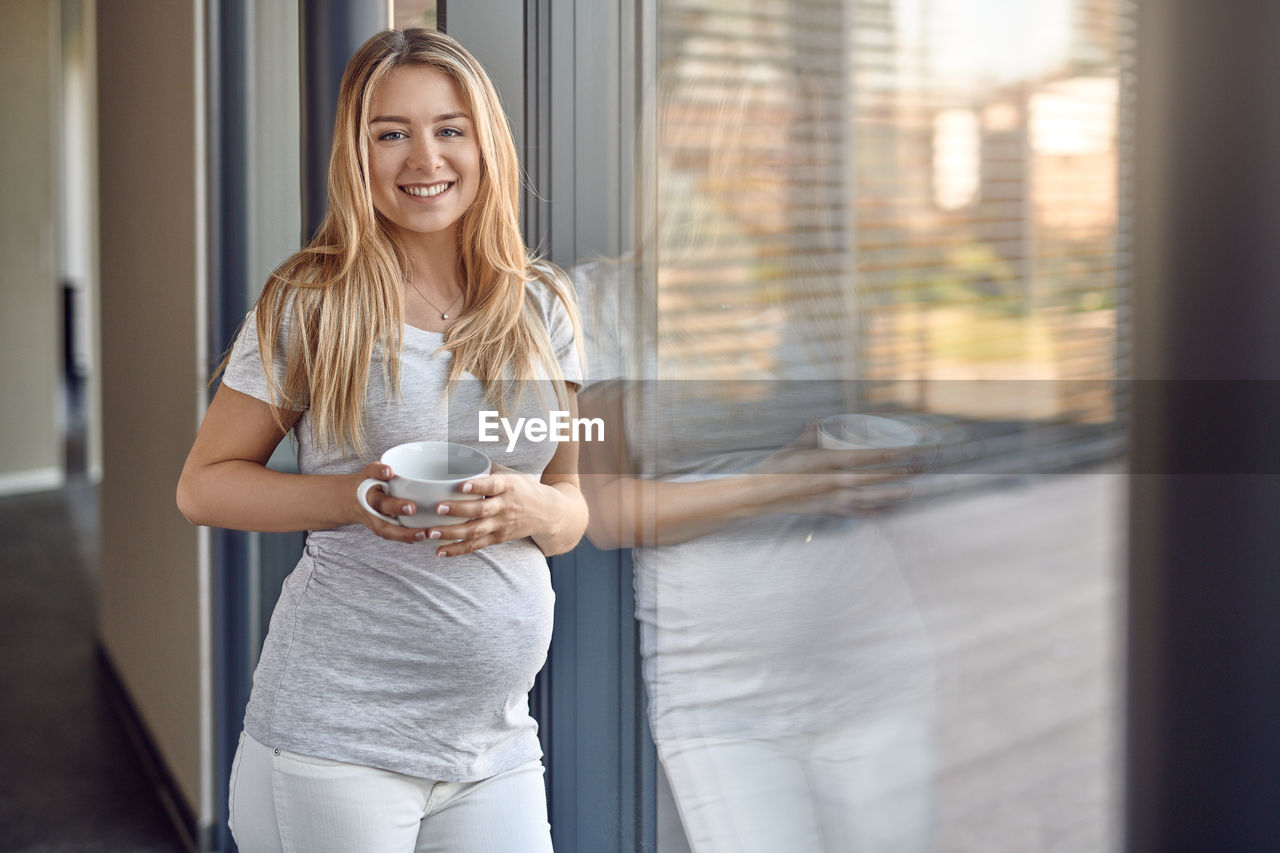 Portrait of smiling pregnant young woman with blond hair holding coffee cup by window at home