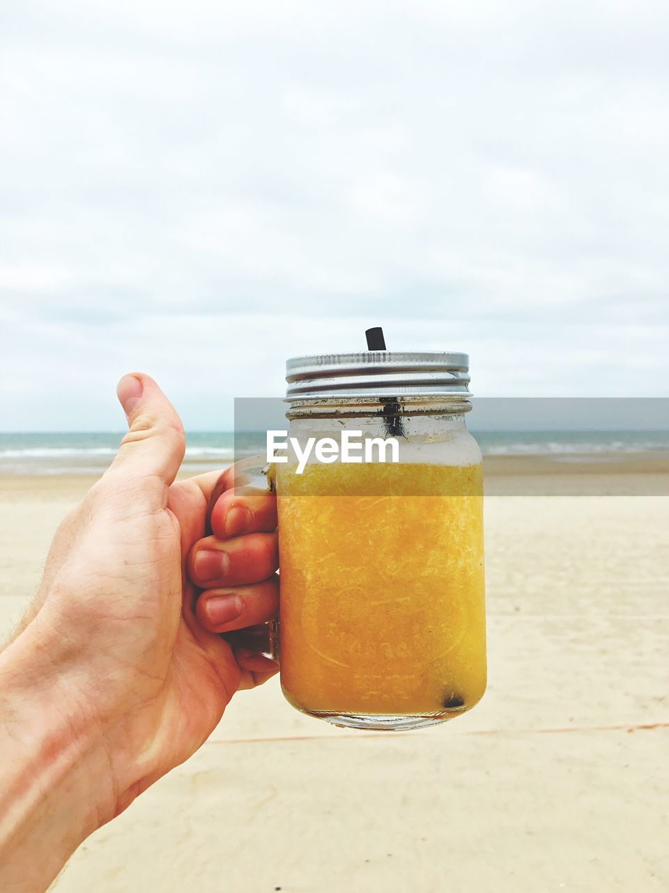 Cropped image of hand holding drink in mason jar at beach against sky