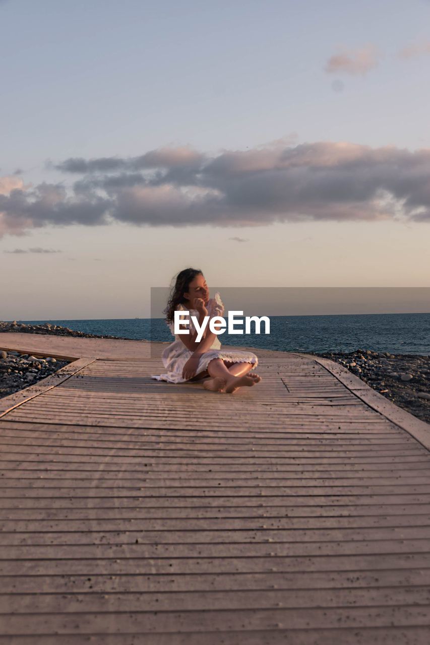 Full length of girl sitting on boardwalk at beach against sky
