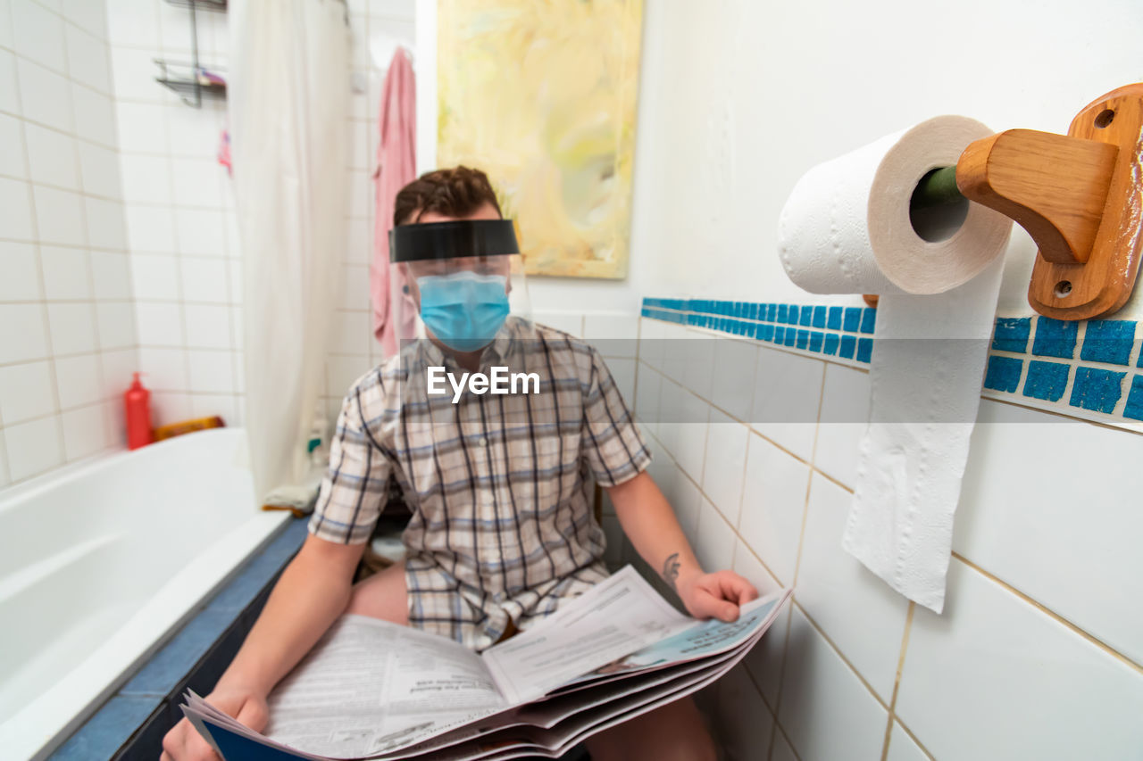 Man wearing mask reading newspaper while sitting in bathroom