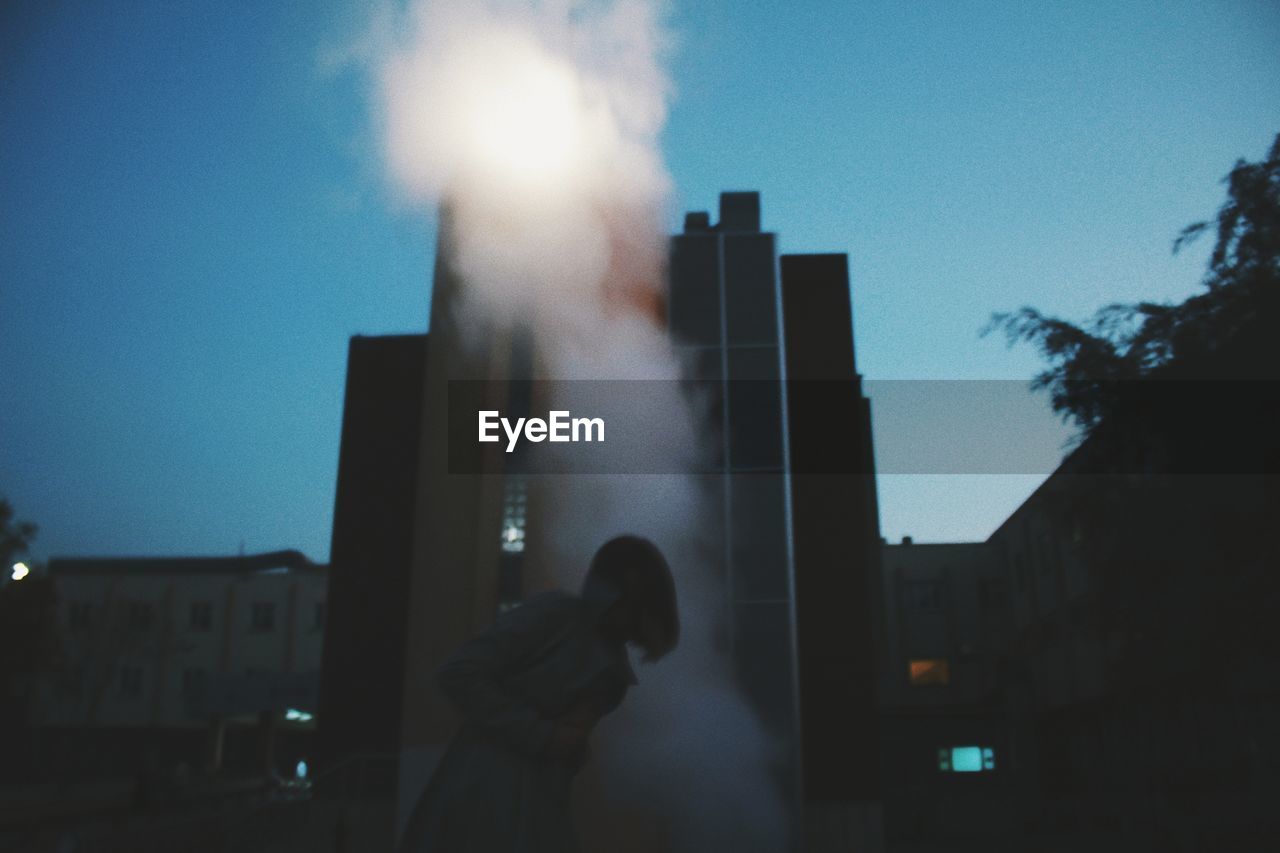 Silhouette woman standing in smoke against buildings at dusk