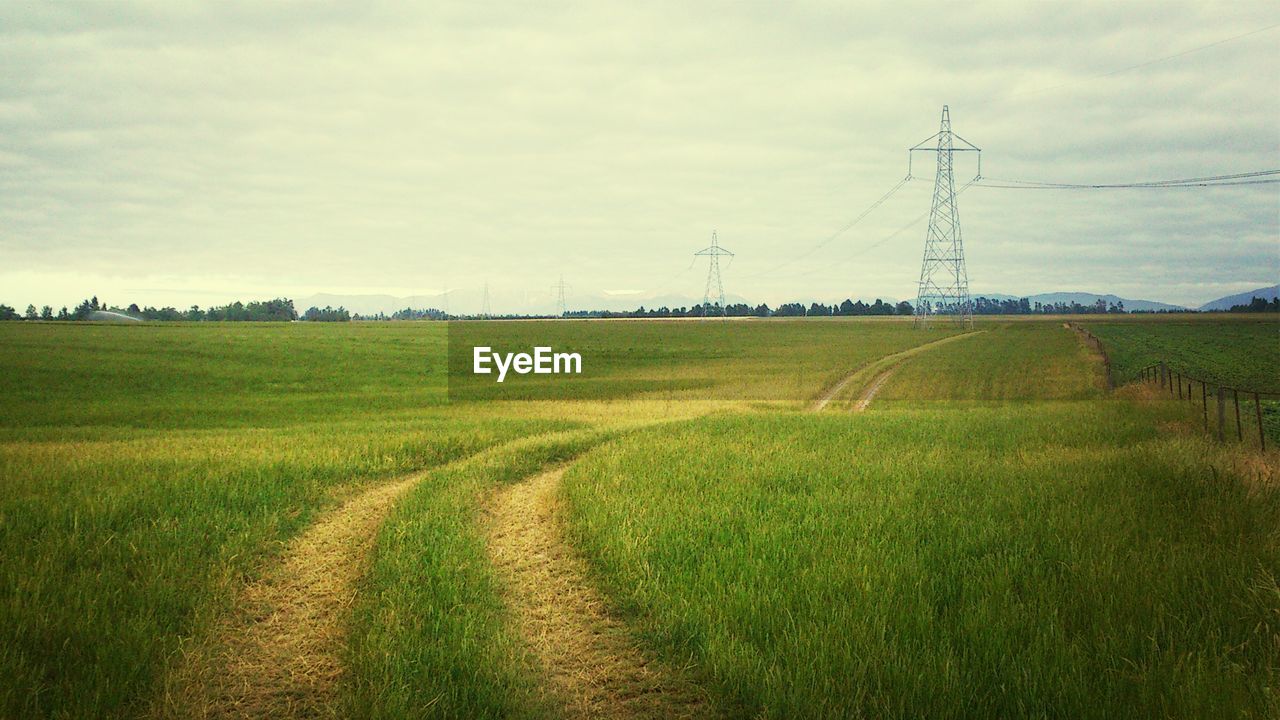 Electricity pylons on grassy field against cloudy sky