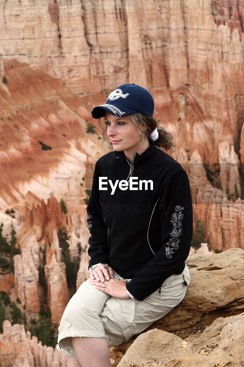Woman wearing cap looking away while sitting on cliff against dramatic landscape