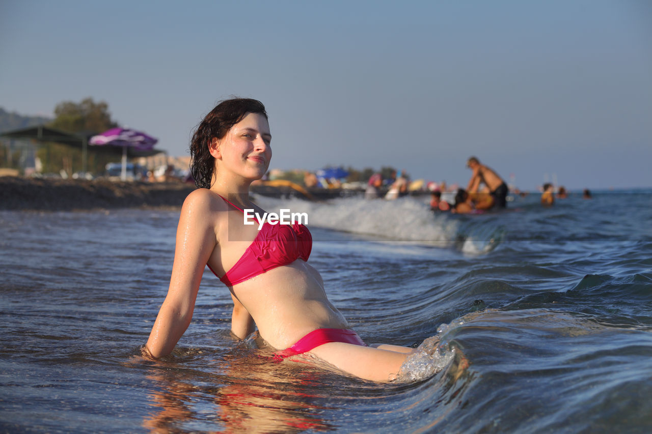 Side view of young woman sitting on shore at beach against clear sky