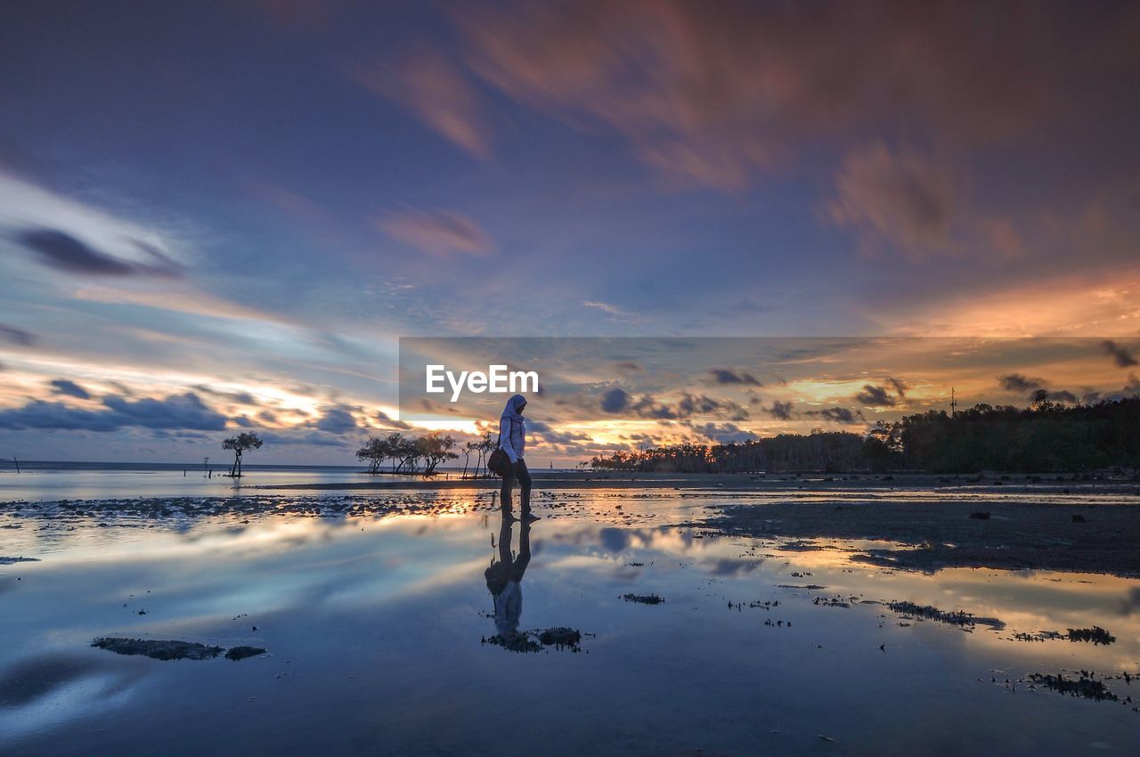 WOMAN STANDING ON BEACH AGAINST SKY DURING SUNSET