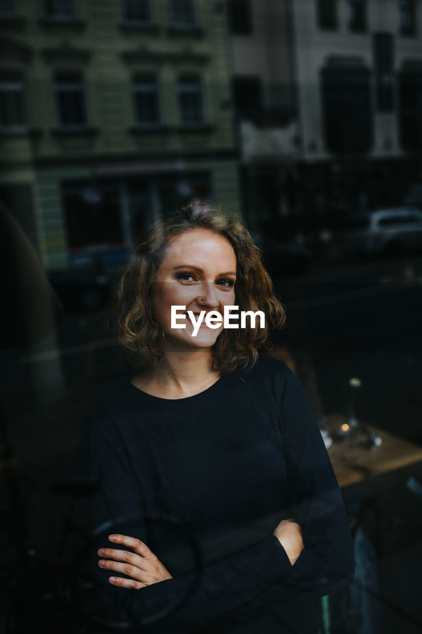 Portrait of smiling young woman with arms crossed seen through glass window