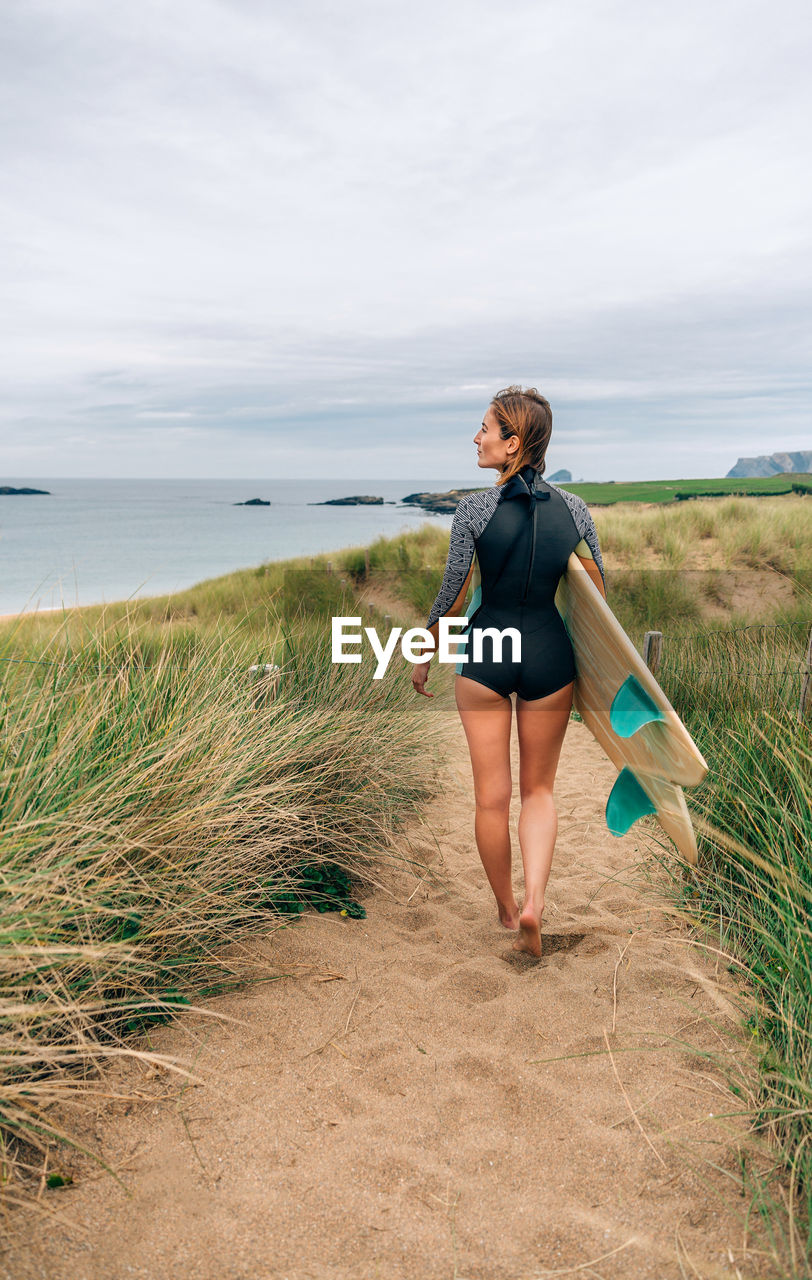 Surfer woman with wetsuit and surfboard going to the beach through the dunes