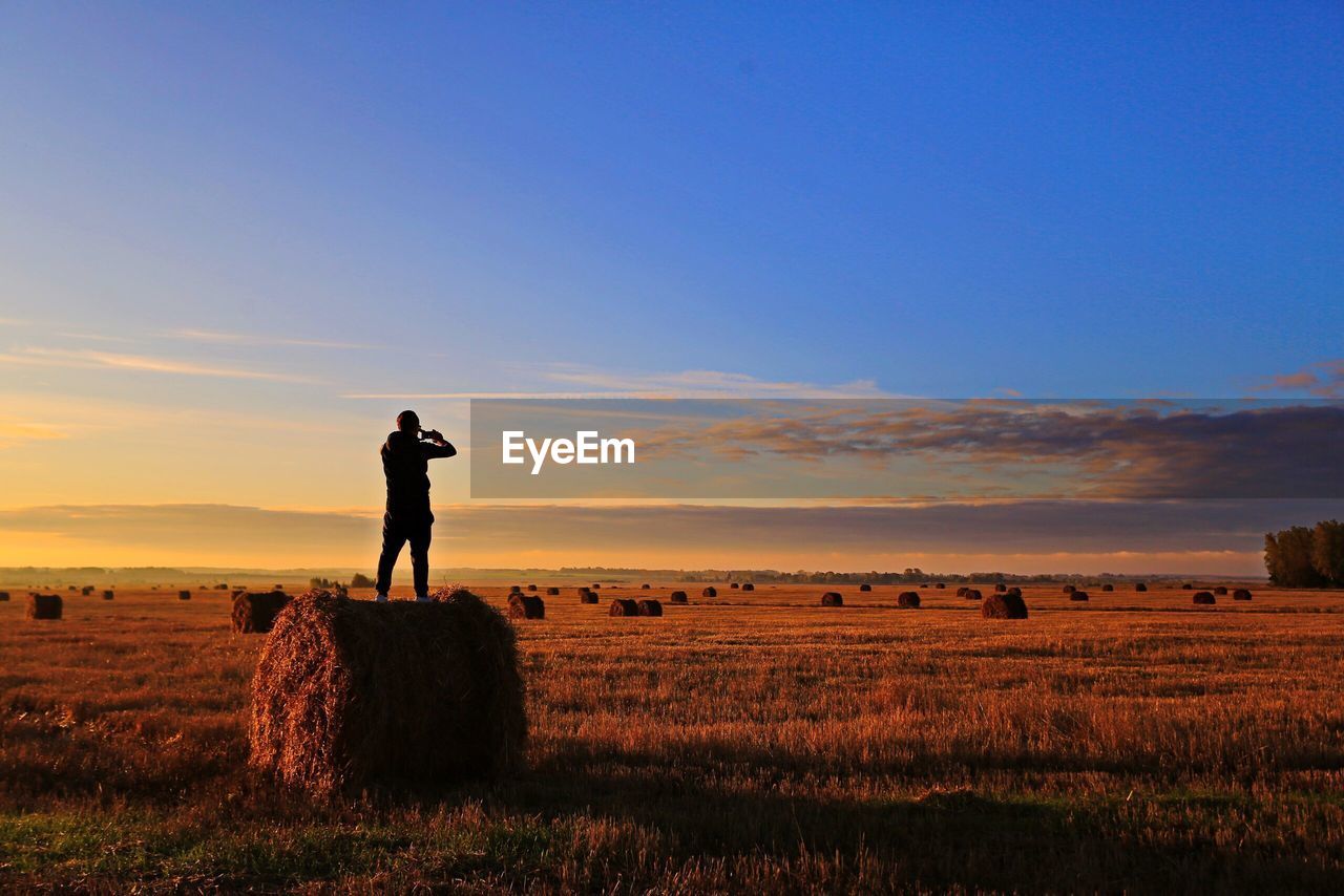 Man standing on hay bale at farm against sky during sunset
