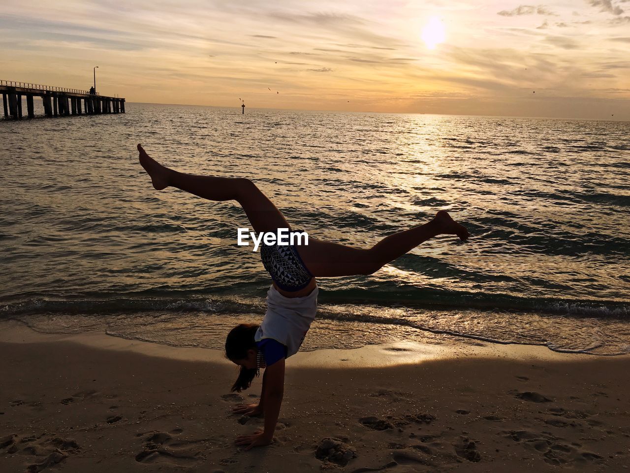 Full length of woman perching handstand at beach against sky during sunset