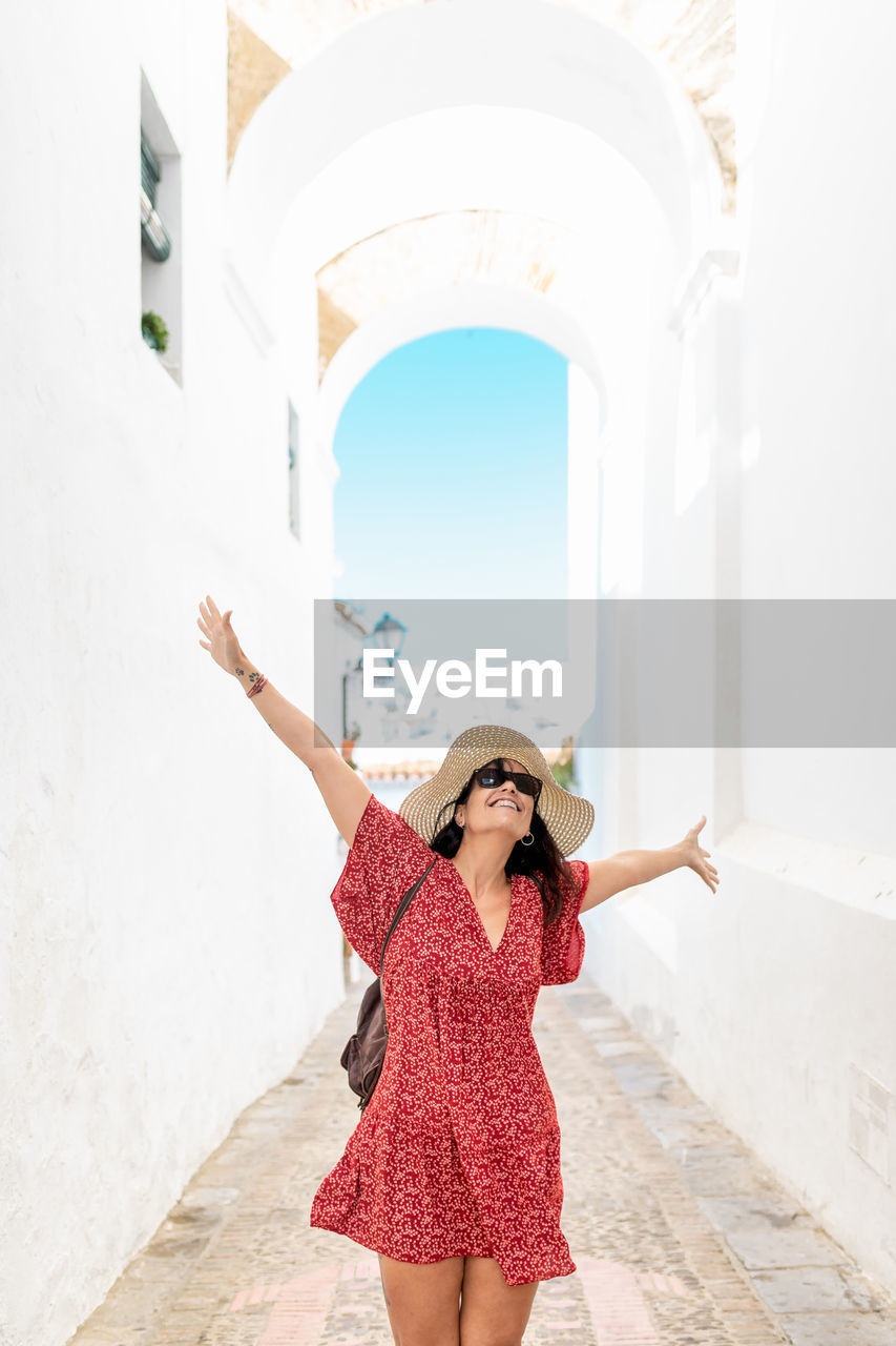 Positive female tourist in straw hat and sunglasses standing with raised arms on narrow street in white village while looking up