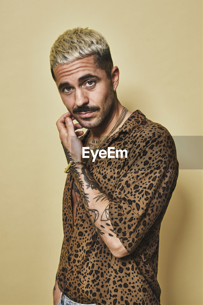 Fashionable male model with tattoos wearing trendy leopard shirt and jeans standing against beige background and looking at camera