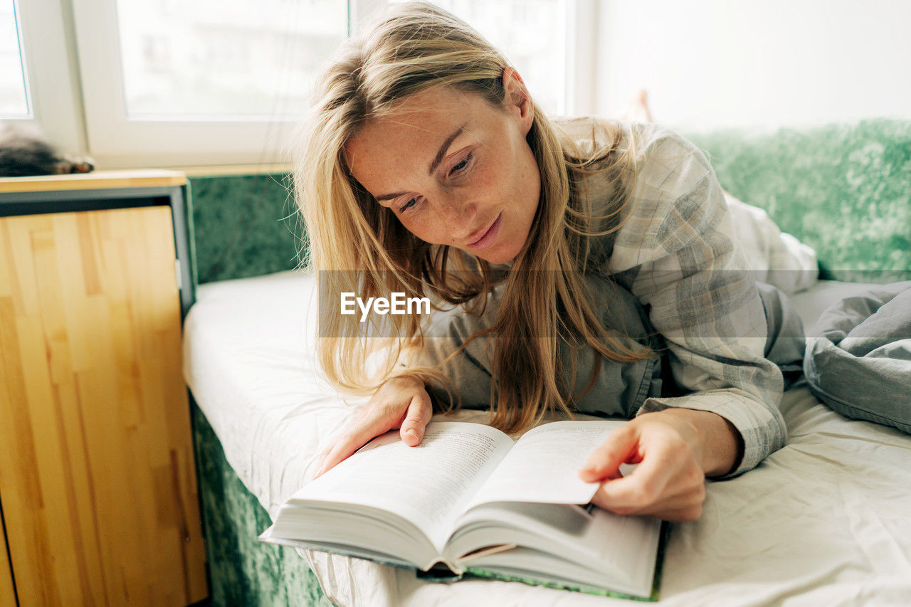 Attractive freckled blond woman in her thirties reading a book while lying in bed. person