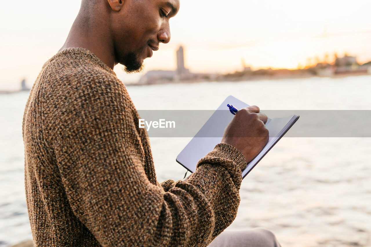 Side view of cropped content african american male taking notes in notepad while sitting on rocky coast near sea in city