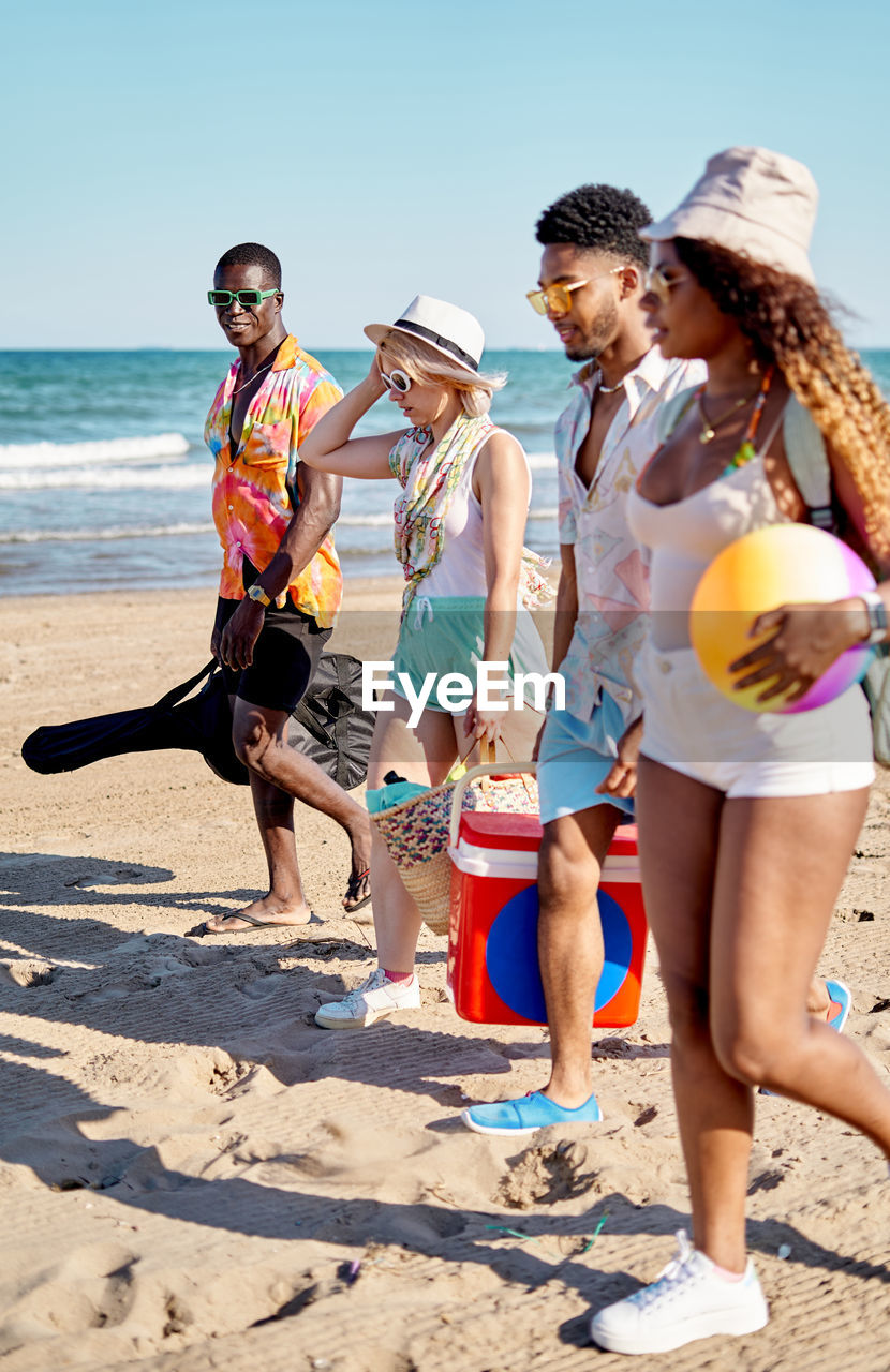 Full body side view of multiracial men and women in sunglasses and hat strolling on sand against cloudless blue sky while spending time on beach on summer day