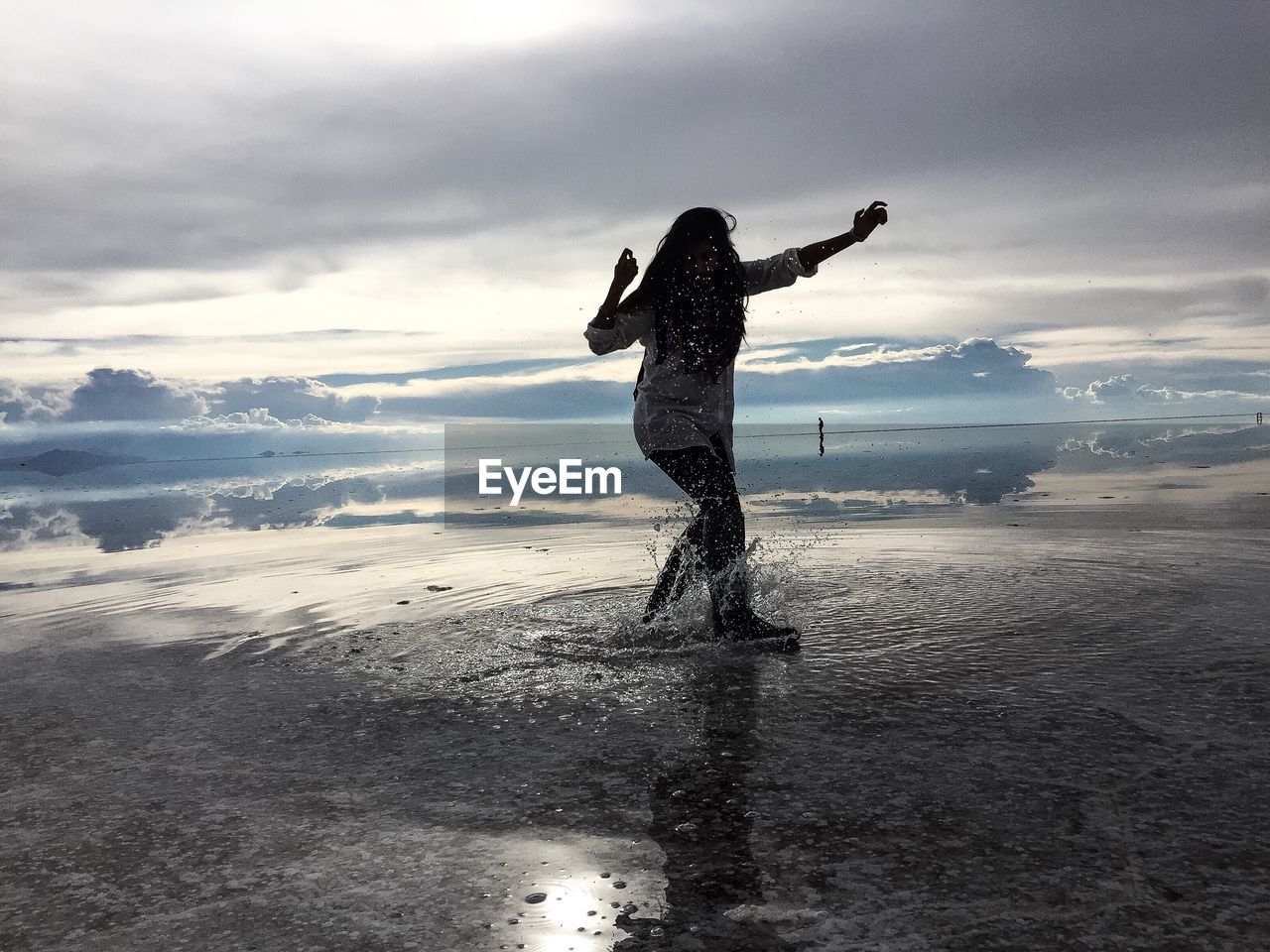 Young woman splashing water at beach against sky during sunset
