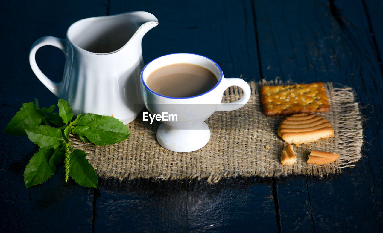 A cup of tea with tasty biscuits, teapot and fresh leaves on old wooden dark background