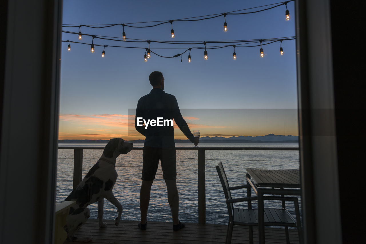 Silhouette man with dog standing by railing against sea during sunset